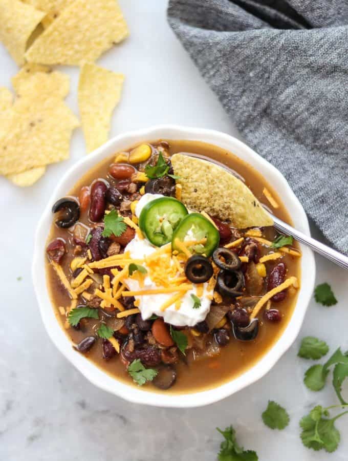 overhead photo of vegan taco soup in white bowl with a variety of vegan garnishes including vegan sour cream and cheese