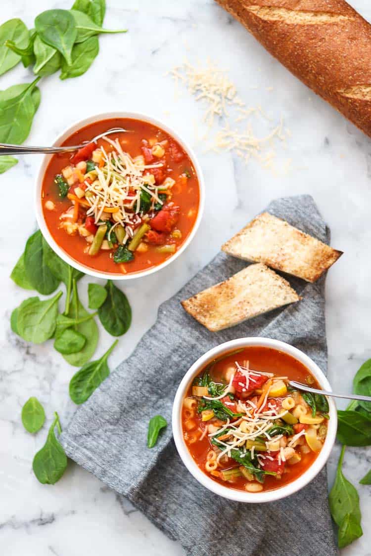 wide overhead shot showing two bowl of vegan minestrone with napkins, bread, and spinach on a marble background