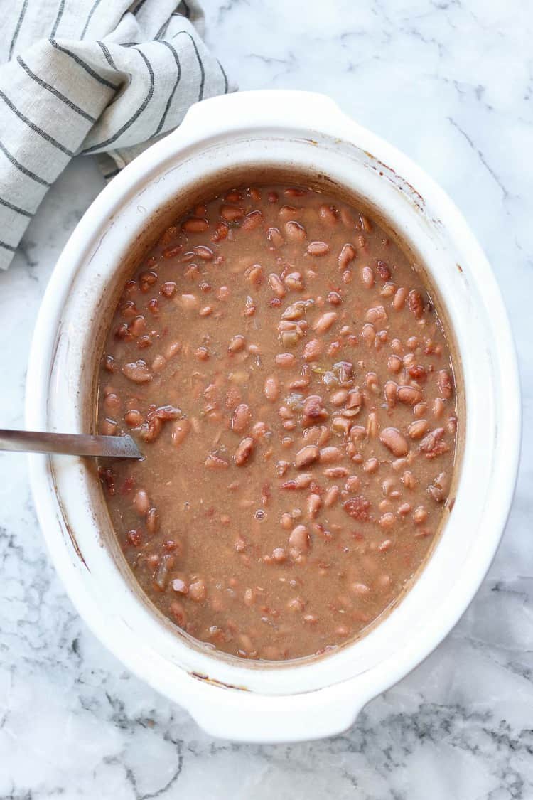 overhead photo of refried beans in a white crockpot with a silver ladle on a marble background