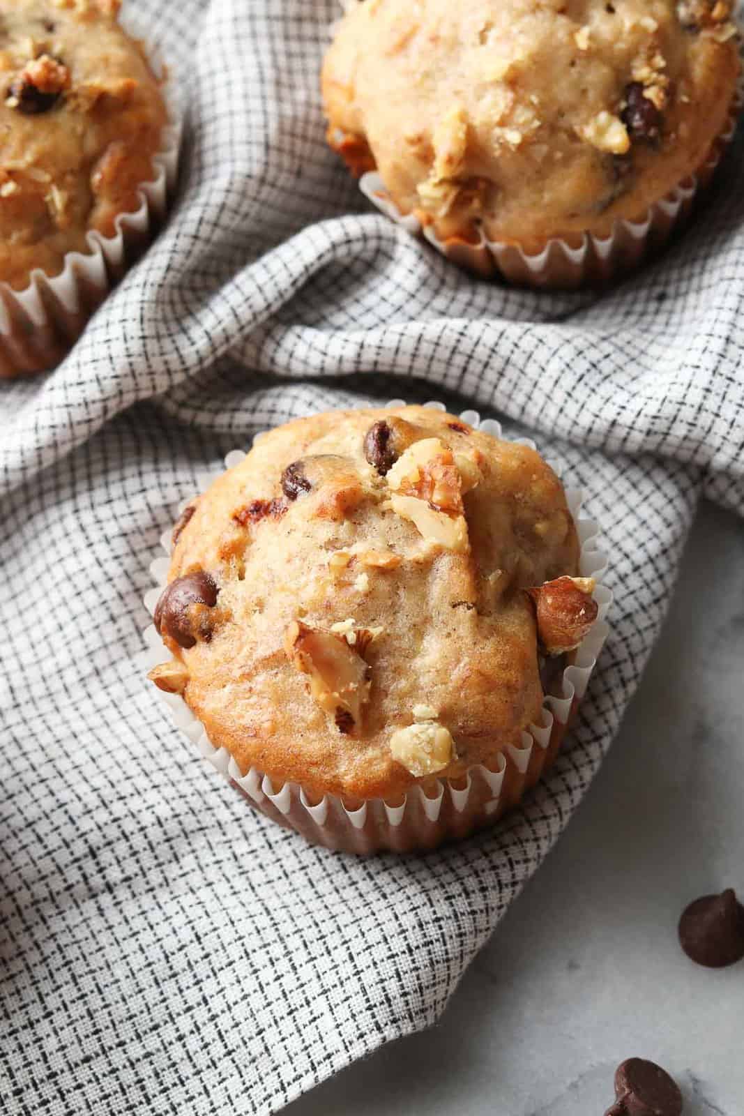 side overhead shot of muffin in white wrapper setting on checked napkin with chocolate chips beside 