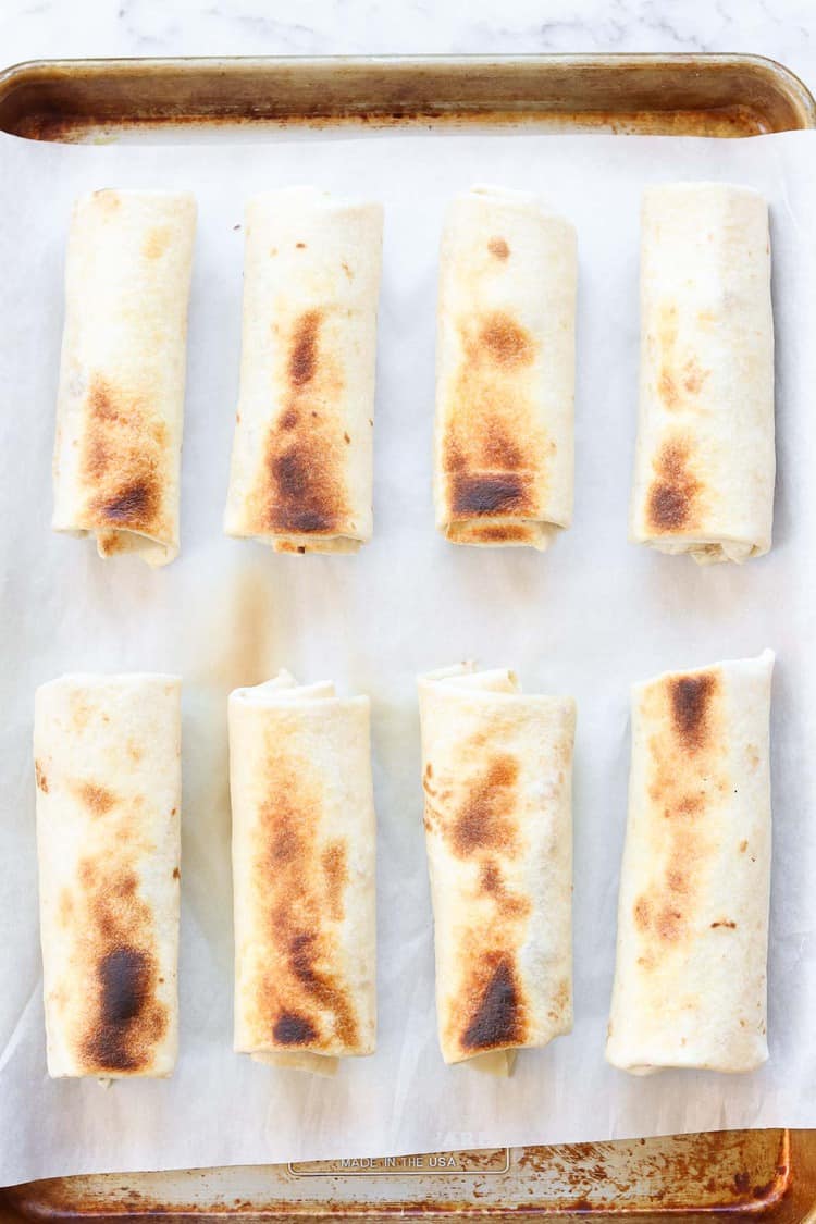 overhead photo of 8 lightly browned baked burritos lined up on a baking sheet 