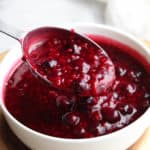 side overhead view of berry sauce in a white bowl with a silver measuring spoon ladling it out