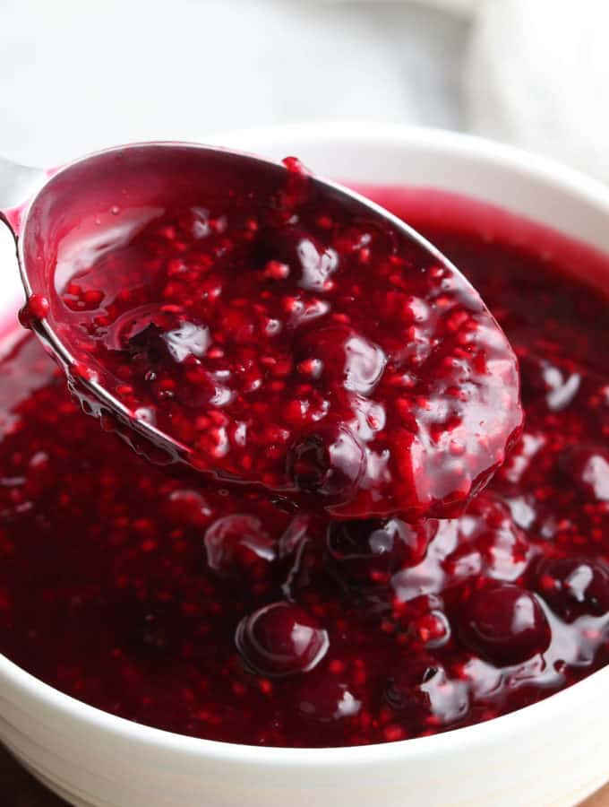 side overhead view of berry sauce in a white bowl with a silver measuring spoon ladling it out