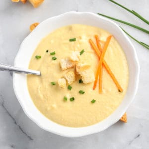 overhead photo of creamy soup in a white bowl garnished with broken croutons, carrot sticks, and chopped chives