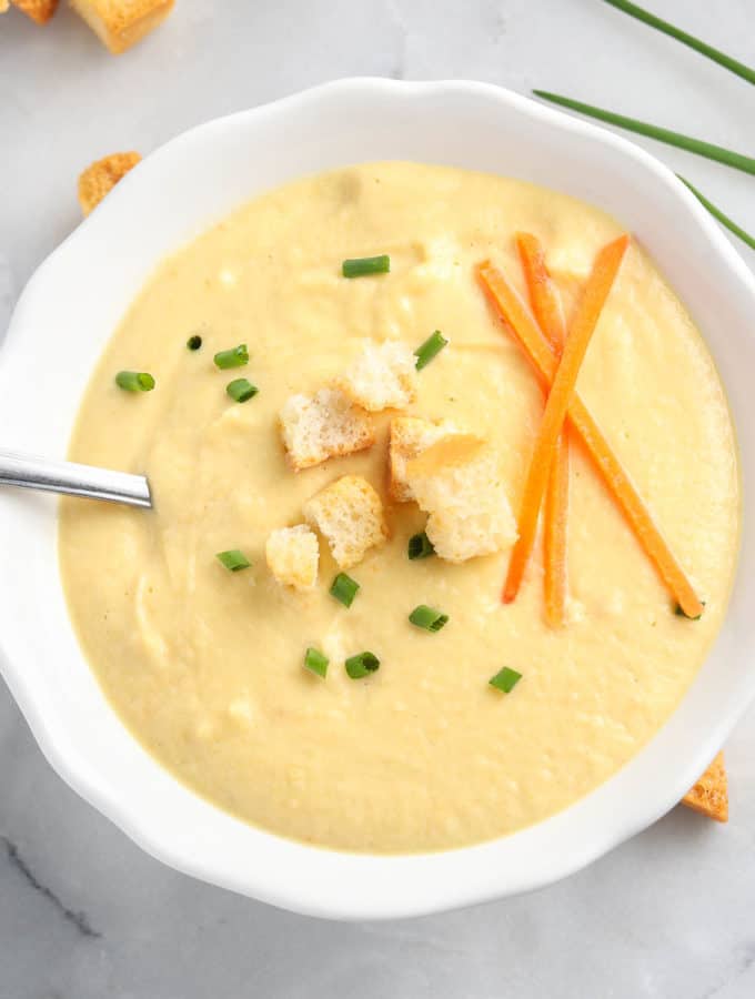 overhead photo of creamy soup in a white bowl garnished with broken croutons, carrot sticks, and chopped chives