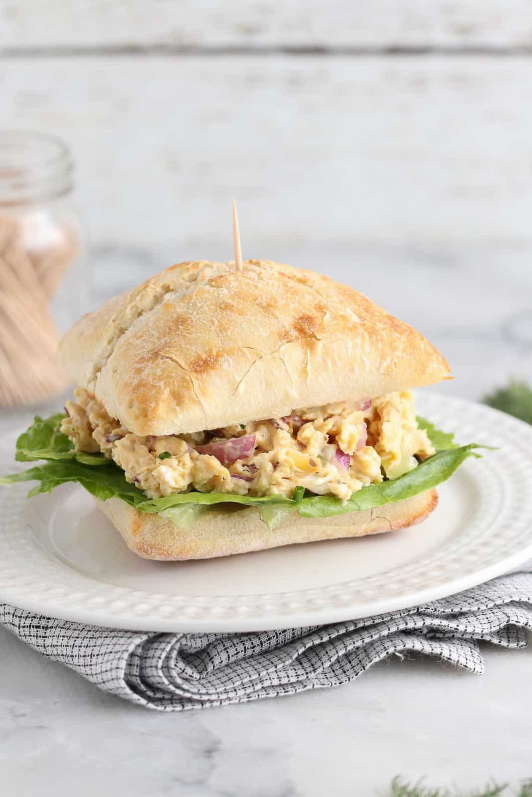 side shot of finished vegan chicken salad in a square roll on a white plate with a toothpick inserted and more in the background