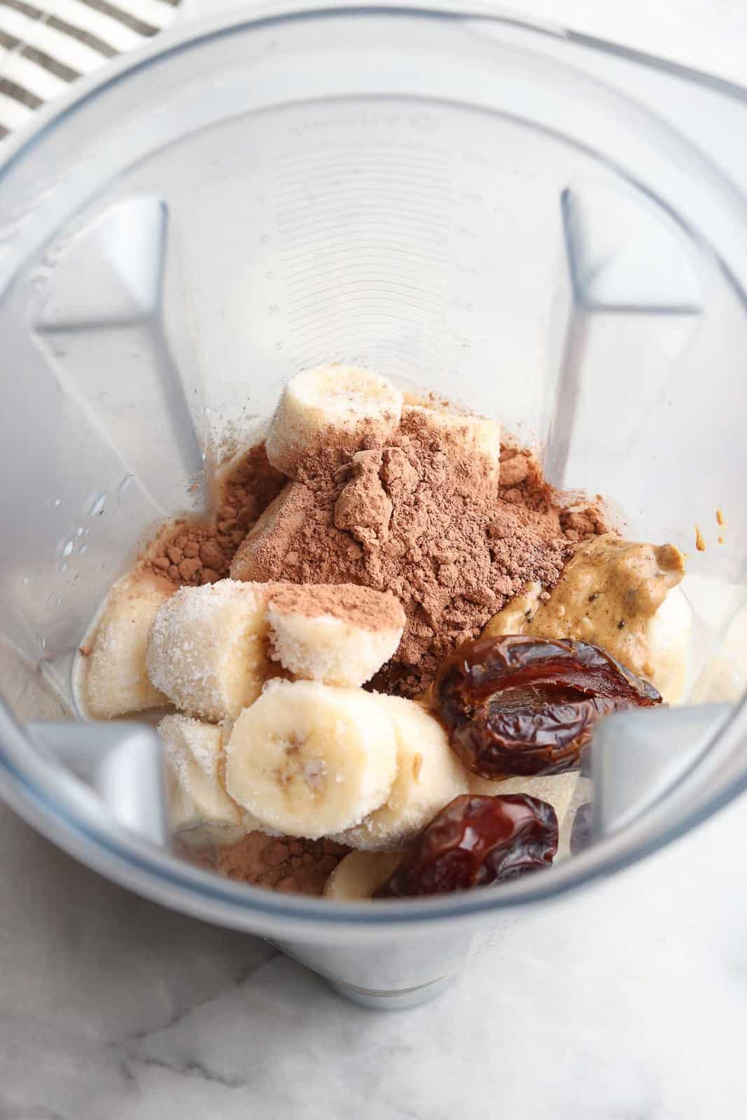 overhead view of blender contents showing banana chunks, cocoa powder, dates, and nut butter