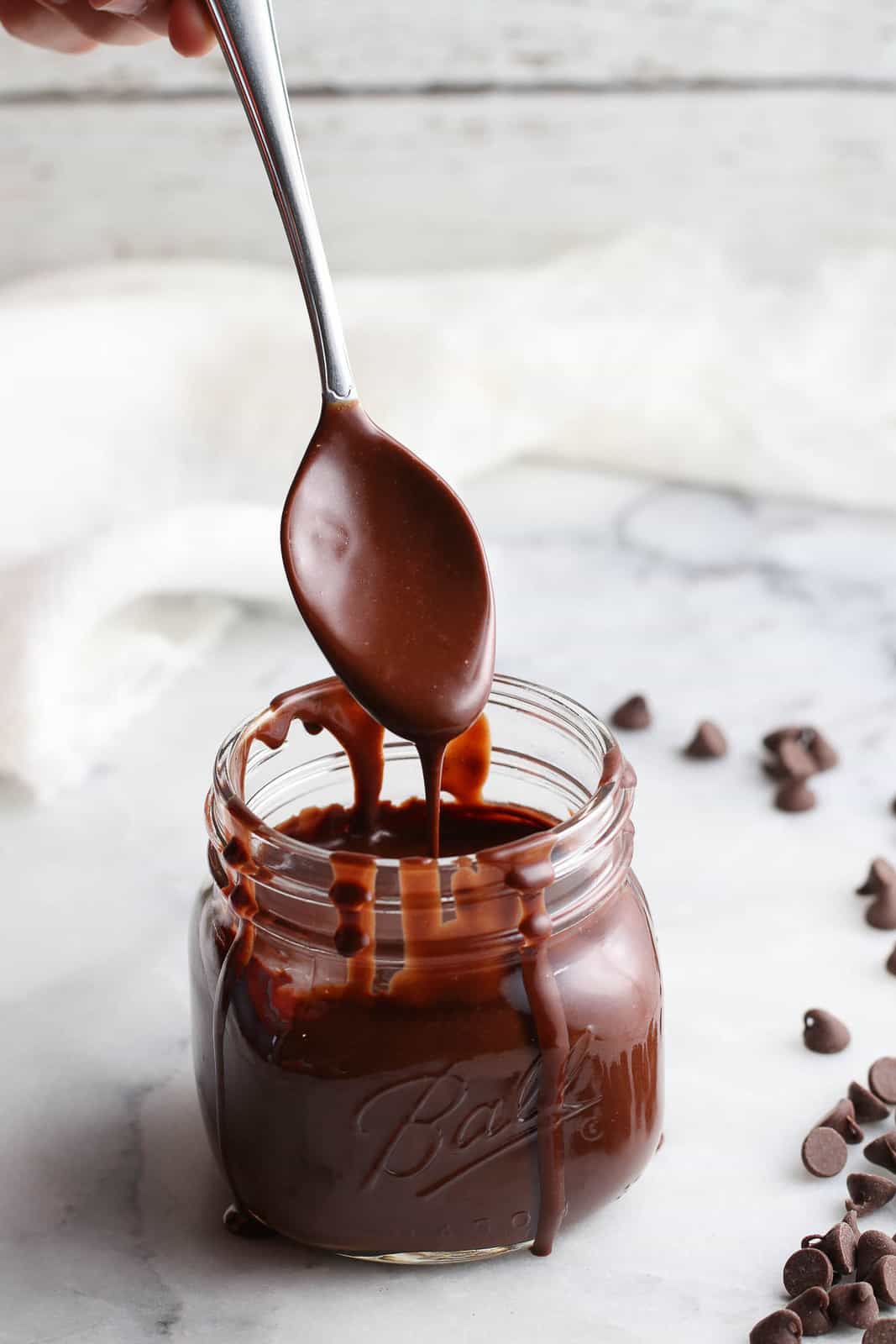 side photo showing silver spoon dripping chocolate sauce into a clear mason jar on a marble background