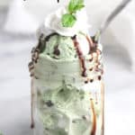 side shot of scoops of mint chocolate chip ice cream in a small mason jar topped with whipped cream, mint sprig, and chocolate drizzle