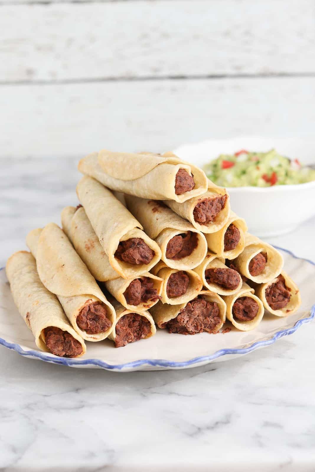 side overhead view of vegan black bean taquitos stacked on a white plate with guacamole in the background