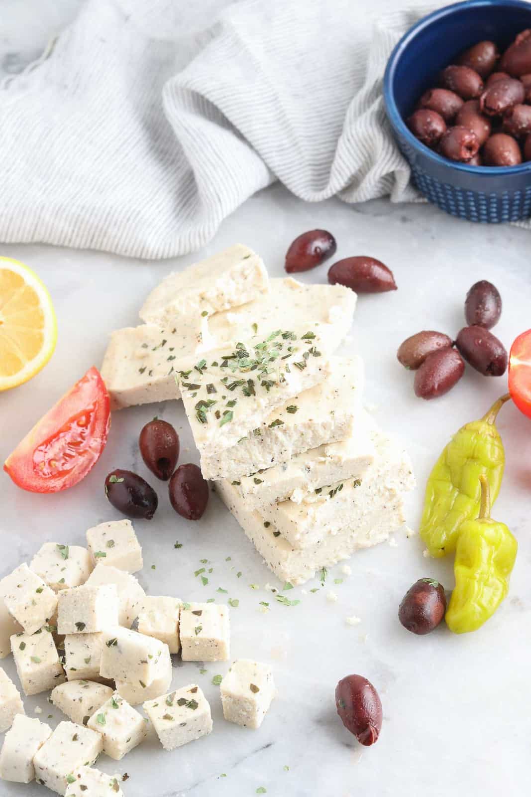 wide overhead photo showing vegan feta cubes and slabs, olives, sliced lemon, wedged tomato and pickled peppers