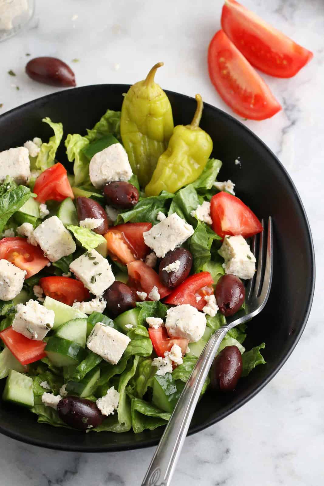 overhead view of chopped green salad with diced tomatoes, greek olives and pickled banana peppers and crumbled vegan feta in a black dish