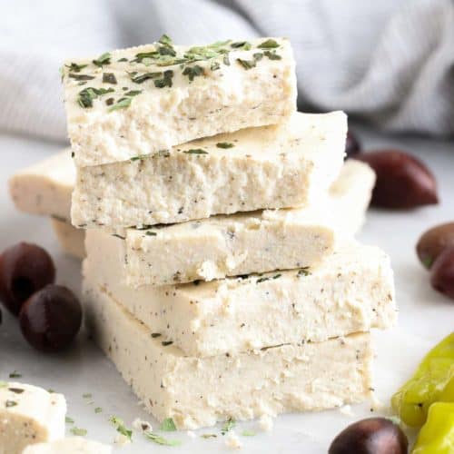 side view of squares of vegan feta stacked on each other with greek olives and peppers nearby