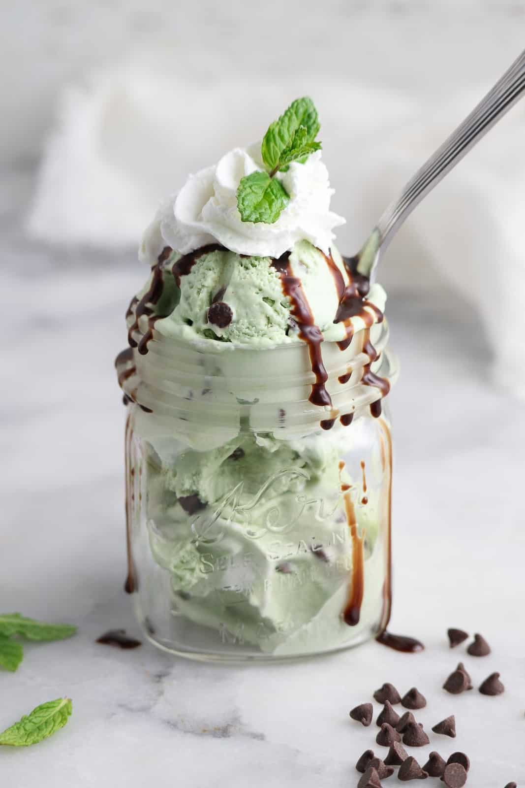 side view of mint chip ice cream scooped into small mason jar with drizzle of chocolate syrup and topped with whipped cream and a sprig of mint.