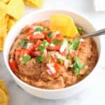 overhead view of white bowl filled with refried bean dip and topped with pico salsa with corn chips beside