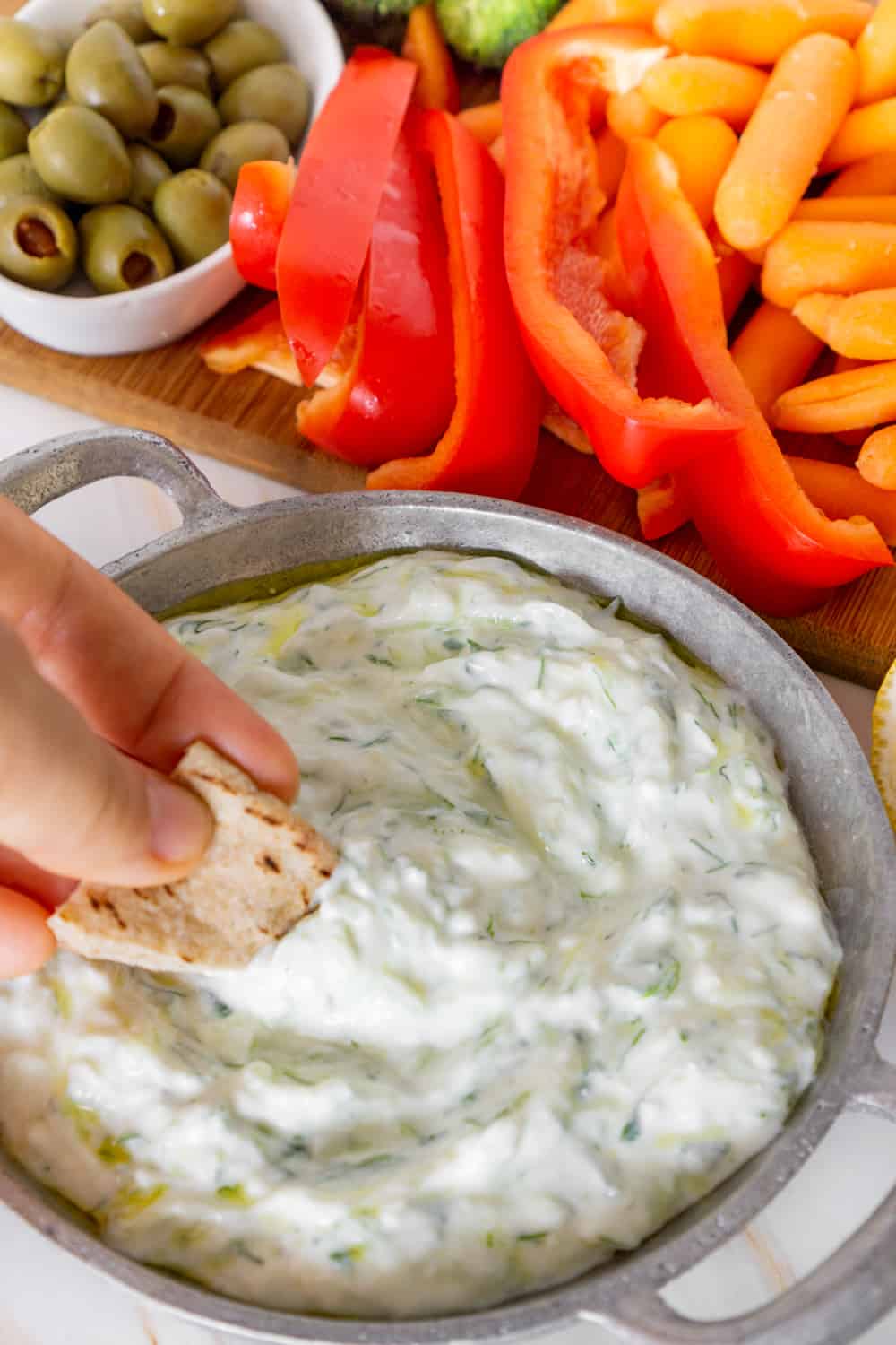 hand dipping pita chips into a dairy free tzatziki dip with crudités in the background