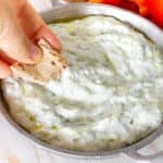 hand dipping pita chips into a dairy free tzatziki dip with crudités in the background
