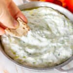 hand dipping pita chips into a dairy free tzatziki dip with crudités in the background
