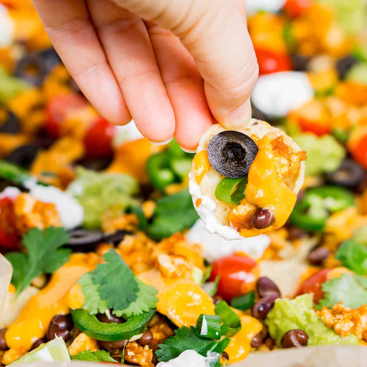 Hand holding a loaded nacho chip with vegan toppings: crispy tofu crumble, vegan nacho cheese, olives, vegan sour cream
